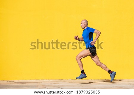 Photo with copy space of an adult man with shaved head and sportswear running in the street in front of a yellow wall