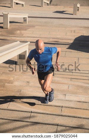 Vertical photo of the adult male with shaved head and sportswear running up stairs in a park on a sunny day