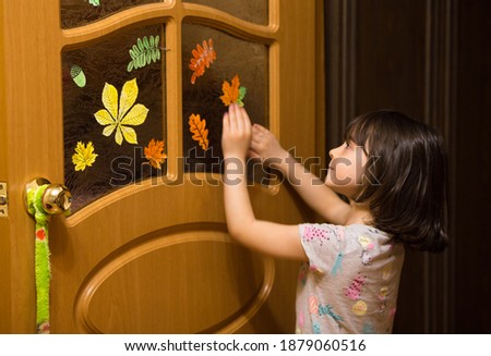 kid girl decorates the door in the room with paper pictures of autumn leaves. maple, rowan, birch colored paper decorations.