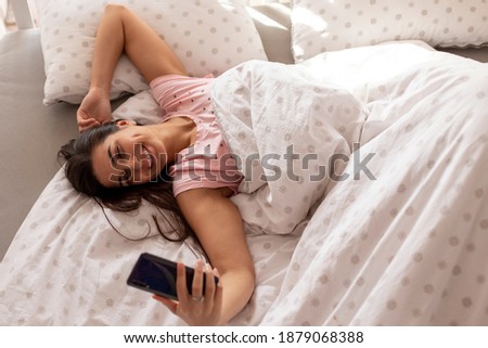 A smiling young woman is taking a selfie on her mobile phone while lying in bed in the morning.