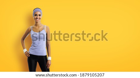 Portrait image of smiling sporty brunette woman in sportswear, isolated over yellow background. Young female fitness instructor or personal trainer at studio.
