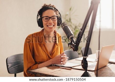 Cheerful woman recording a podcast from home. Female wearing headphones sitting at table with laptop and microphone looking at camera and smiling.