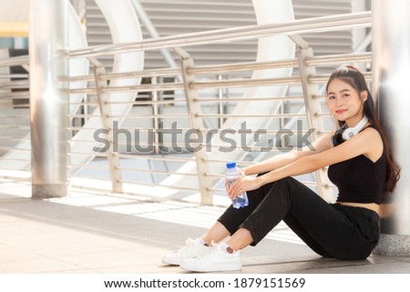 Portrait happy beautiful young girl athlete with black sporty clothes and white headphone sitting and Relaxing after morning training on walkway. People, sport and relaxation concept.