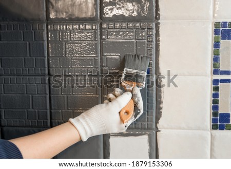 Repainting old dated kitchen ceramic tile back wall with modern gray chalk paint indoors at home. Giving old kitchen new look concept. Hand holding a paint brush tool. Royalty-Free Stock Photo #1879153306