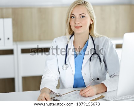 Portrate of woman-doctor at work while sitting at the desk in clinic. Blonde cheerful physician filling up medical form or prescription