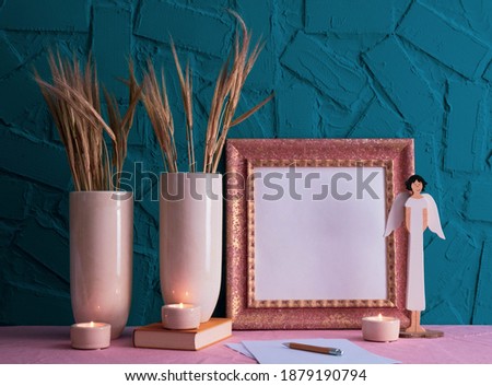 an old book, white vases with ears of corn, burning candles, an angel figure and an antique golden photo frame stand on the table, on a blue background