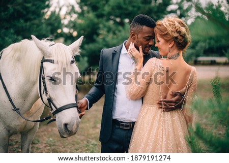 Interracial wedding. African man and Caucasian woman stand embracing near a white horse.