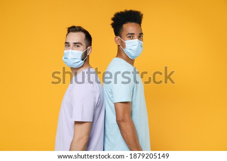 Side view of young friends european african american men in purple blue t-shirts sterile face mask to safe from coronavirus virus covid-19 looking camera isolated on yellow background studio portrait