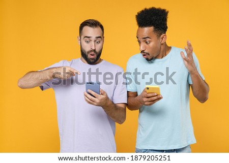 Shocked young two friends european african american men 20s in violet blue casual t-shirts pointing index finger on mobile cell phone typing sms message isolated on yellow background studio portrait