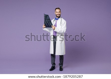 Full length of smiling young bearded doctor man in medical gown hold x-ray brain by radiographic image ct scan mri isolated on violet background studio. Healthcare personnel health medicine concept Royalty-Free Stock Photo #1879205287