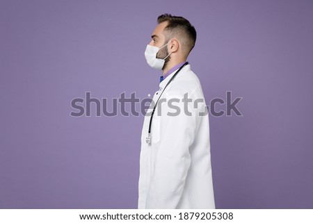 Side view of young bearded doctor man in medical gown stethoscope sterile face mask to safe from coronavirus virus covid-19 isolated on violet background. Healthcare personnel health medicine concept Royalty-Free Stock Photo #1879205308