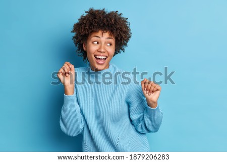 Photo of joyful dark skinned woman dances carefree keeps fists raised looks positively aside dressed in casual jumper moves against blue background. People positive emotions and feelings concept