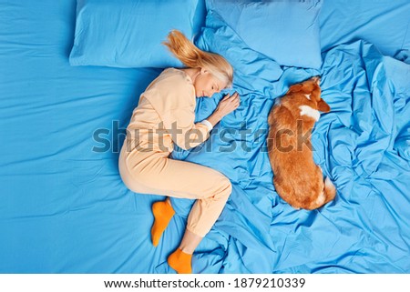 Above view of senior woman in nightwear has good deep sleep with dog poses on blue bedclothes lies in favorite pose for rest sees pleasant dreams. Asleep female at comfortable bed favorite pet near