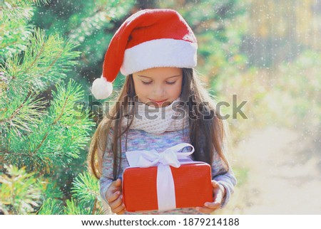 Christmas portrait of happy smiling little girl child in santa red hat with gift box near a green branch tree