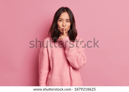 Serious mysterious brunette Asian woman presses index finger to lips makes hush gesture tells secret asks to be quiet wears long sleeved jumper poses against pink background. Secrecy concept Royalty-Free Stock Photo #1879218625