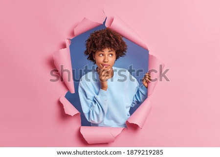 Dark skinned female model keeps hand near mouth as if trying to remember something looks thoughtfully above dressed in casual blue jumper breaks through paper background stands in ripped hole