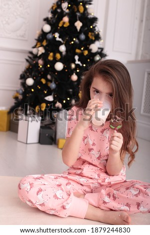 A girl in pink pajamas sits by a New Year tree with a chocolate candy on a stick and a glass of milk Christmas card
