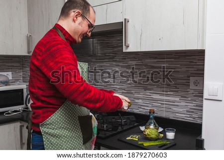 A man in a red fleece and a green apron cooking grilled artichokes and asparagus in a kitchen. He is cutting artichokes.