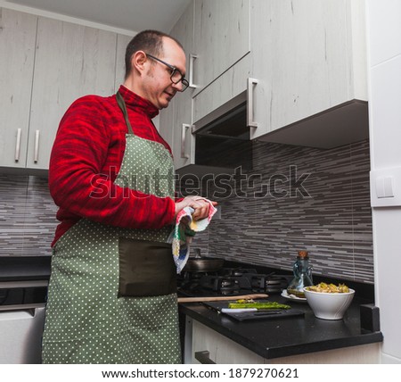 A man in a red fleece and a green apron cooking grilled artichokes and asparagus in a kitchen. He is drying his hands.