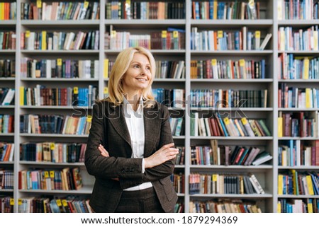 Beautiful mature lady with cross hands stands in modern office. Successful business woman in formal suit, employee or teacher looks away and smiling