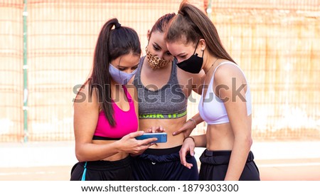 Three cheerful girls wearing masks look at a picture of a mobile phone while practicing basketball on a court in the city