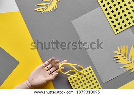 a wooden hand holds a paper bag, the whole composition is set on a grey and yellow geometric background. Illuminating Yellow, Ultimate Grey, 2021 colors. Trendy monochrome image.
