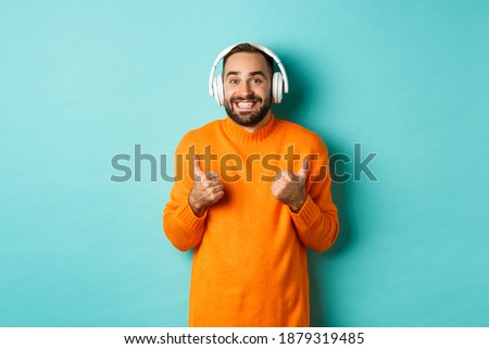 Amazed adult man listening music in headphones, looking at camera impressed and showing thumbs-up in approval, recommending, standing over turquoise background
