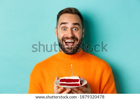 Close-up of happy adult man celebrating birthday, holding bday cake with candle and making wish, standing against turquoise background