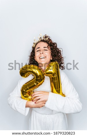 A beautiful brunette woman laughing while hugging two golden balloons with the number twenty-one for the New Year. She is wearing pajamas and a Christmas headband. New Year's Eve at home concept