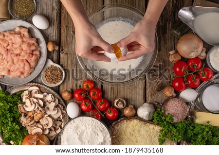 Chef hands breaks egg into glass bowl with milk for preparing dough on wooden table with variety of ingredients background. Concept of cooking process. Backstage of cooking pie. View from above.
