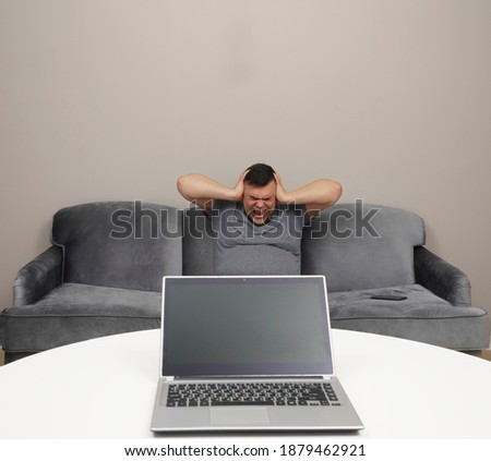 Man Sitting On Sofa In Front Fallen. empty Screen of laptop. Male with his laptop in a room covering ears with hands. Frustrated expression