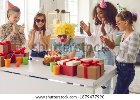 Little boy in clown wig and nose making his birthday wish before blowing candles on cake surrounded by happy diverse children in funny party hats and sunglasses who are clapping hands