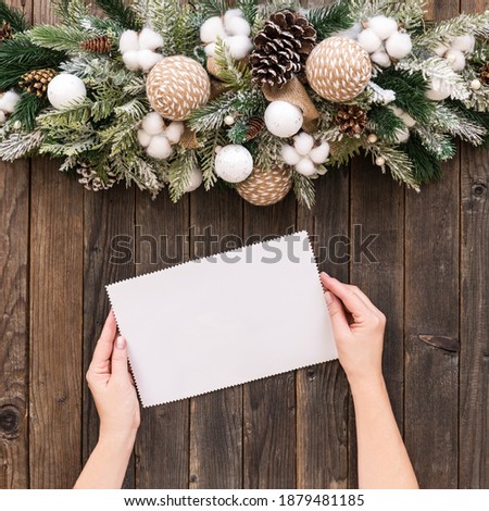 Christmas composition and women hands holding sheet of paper or brochure or invitation on wooden background. Flat lay, top view with copy space.