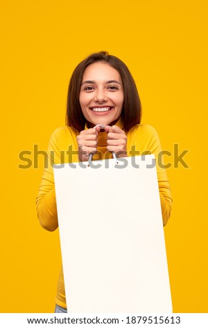 Laughing brunette holding empty white paper bag excited with new purchase looking at camera on yellow background