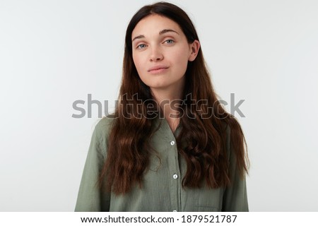 Pleasent looking young brown haired woman with natural makeup keeping her lips folded while standing against white background in green casual shirt