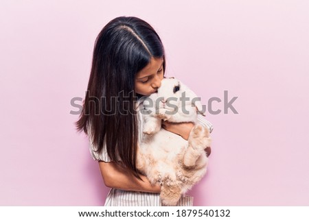 Adorable hispanic kid girl smiling happy. Holding and kissing cute rabbit standing over isolated pink background