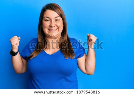 Beautiful brunette plus size woman wearing casual blue t shirt celebrating surprised and amazed for success with arms raised and open eyes. winner concept. 