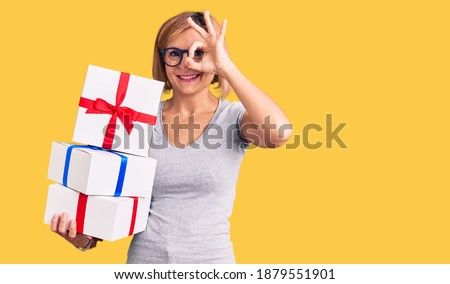 Young blonde woman wearing glasses holding gift smiling happy doing ok sign with hand on eye looking through fingers 