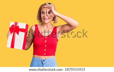 Young blonde woman holding gift smiling happy doing ok sign with hand on eye looking through fingers 