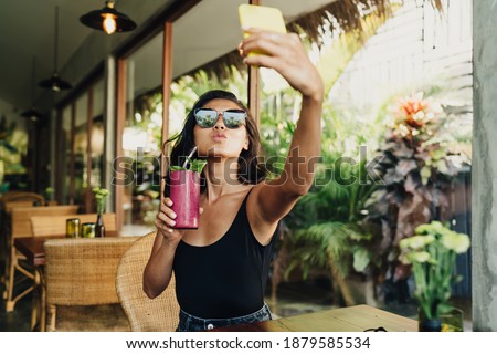 Young female in sunglasses sending air kiss and taking self portrait on blurred background of veranda of modern comfortable restaurant