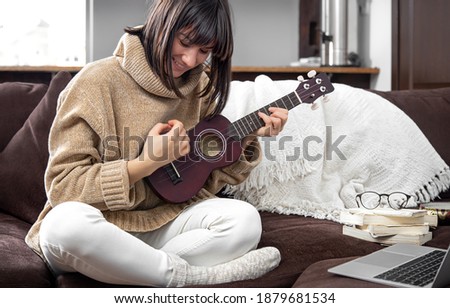 Young cheerful girl in a sweater learns to play the ukulele. The concept of online learning, home education. Royalty-Free Stock Photo #1879681534