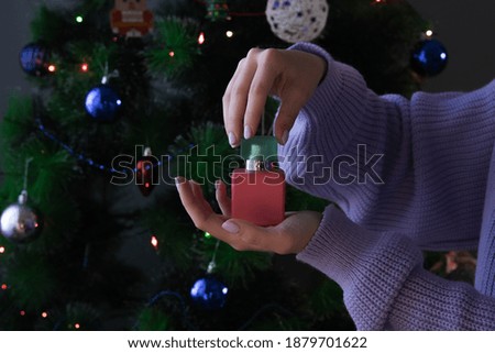 A girl in a purple cardigan holds a bottle of red and green perfume against the background of a Christmas tree with a colorful bokeh.