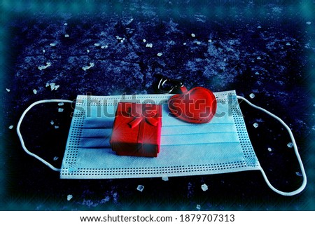 gift box with a red ribbon and a heart on a medical mask on a concrete black background with blue tints. Valentine's day amid covid-19 pandemic.