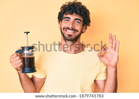 Handsome young man with curly hair and bear holding french coffee maker doing ok sign with fingers, smiling friendly gesturing excellent symbol 