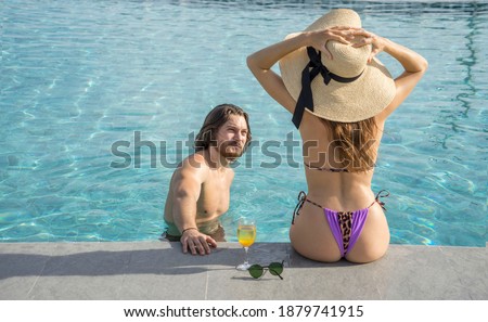 Couple on vacation, a boyfriends swimming, and a girlfriend wearing a bikini, wearing a sun hat, and relaxing at the pool during a honeymoon.