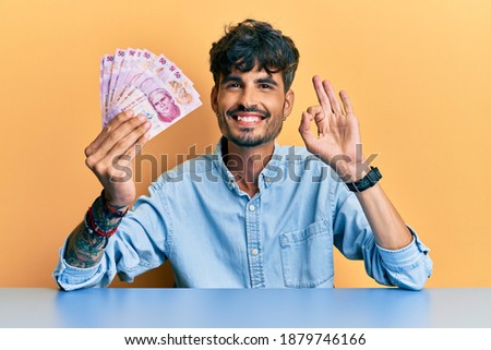 Young hispanic man holding mexican pesos sitting on the table doing ok sign with fingers, smiling friendly gesturing excellent symbol 
