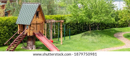 Small wood log playhouse hut with stairs ladder and wooden slide on children playground at park or house yard. Panoramic view. Green grass lawn garden and paved pathway background on bright sunny day Royalty-Free Stock Photo #1879750981