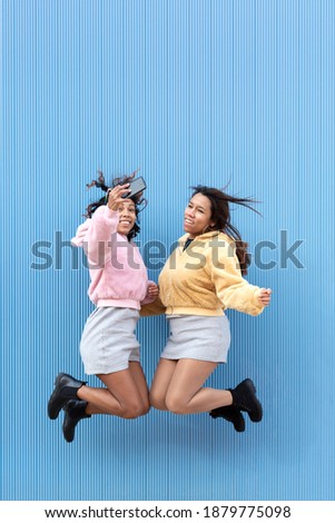 Two happy girls jumping up and down and taking a picture with their mobile phone in the air. Blue background. Space for text.