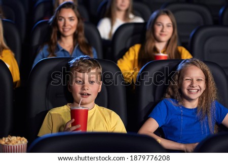 Front view of caucasian kids enjoying time together holding sparkling drink, young audience on background. Happy little friends sitting in cinema, watching funny cartoon. Entertainment concept.