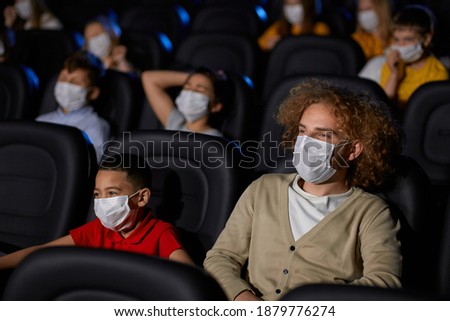 Young man with curly hair enjoying time with little half african brohter during world pandemic. Selective focus of boy watching cartoon in cinema, wearing white face masks, social distancing.